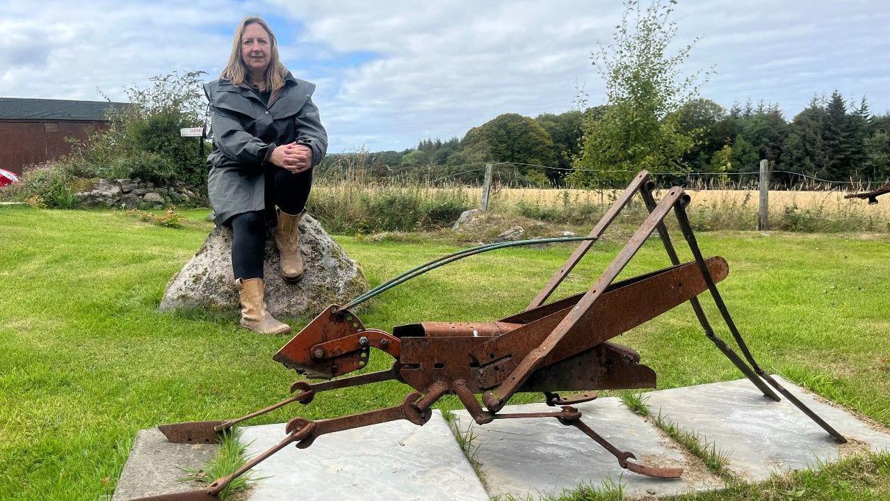 Woman sitting on a rock next to a rusted metal sculpture of a grasshopper. The woman is wearing a long green jacket, dark trousers and brown boots. 
