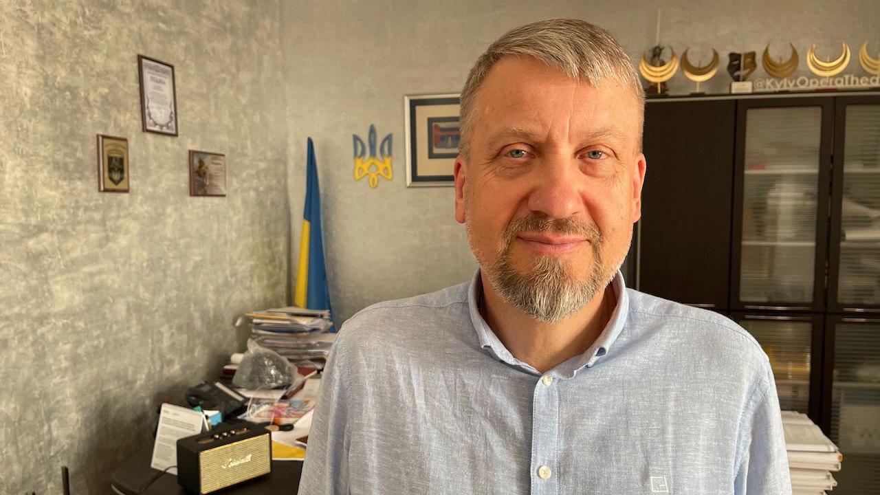 Petro Kachanov is a man in his middle age. He is wearing a light shirt and smiling to the camera. The photo is taken in his office where the Ukrainian coat of arms is hung on the wall.  
