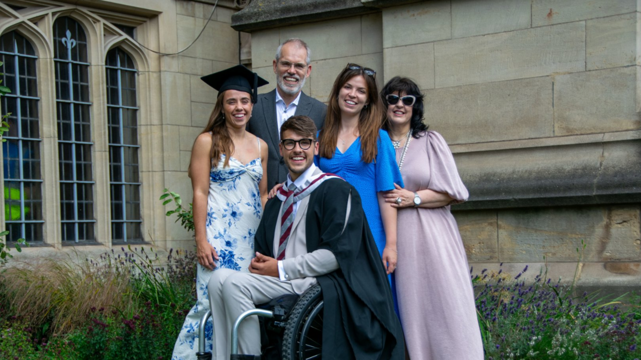 Xander in his graduation robes, sat in a wheelchair, with his wife and three family members around him, smiling at the camera, outside a University of Bristol building