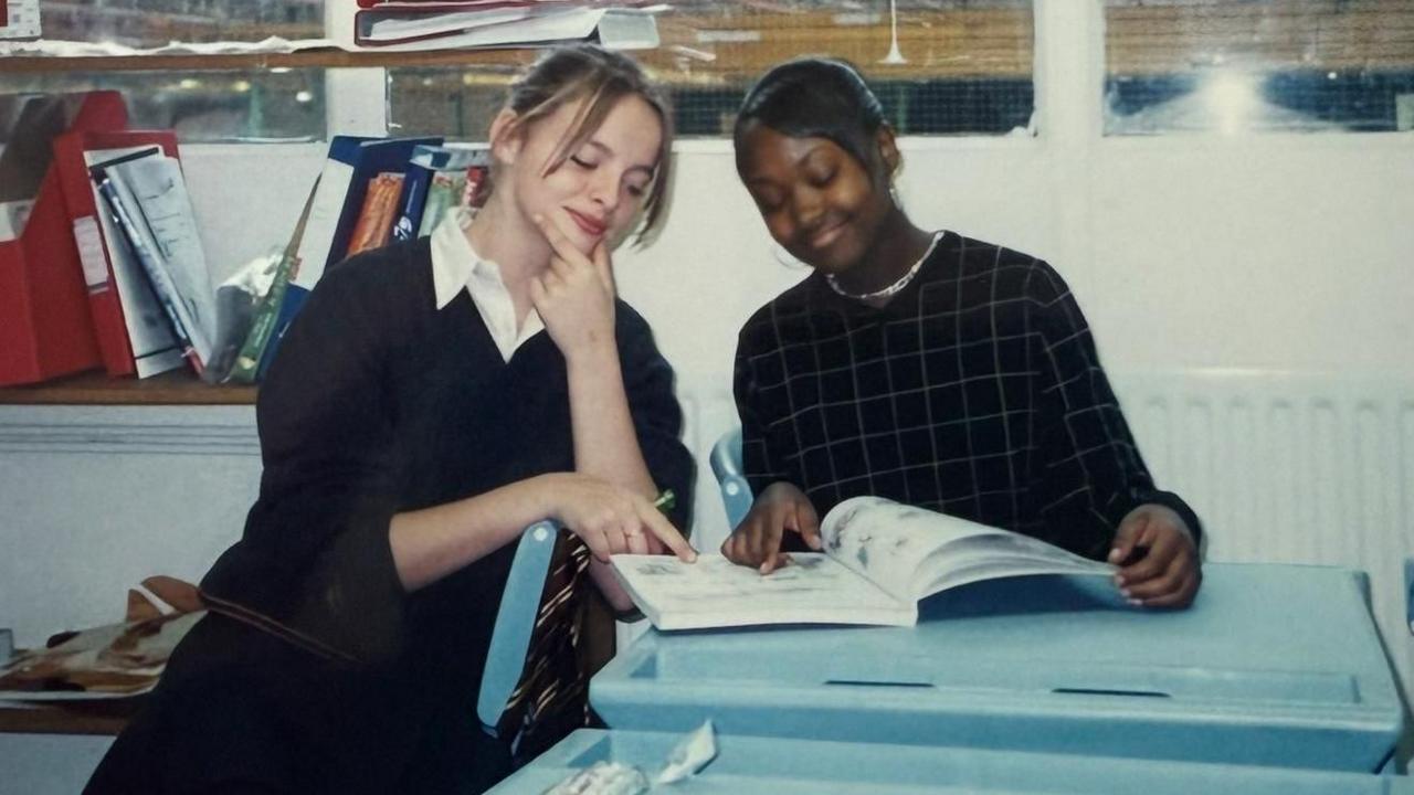 Two people sit at a desk in a classroom looking at an open book. One points at the book. Shelves with books and folders are behind them, and large windows.
