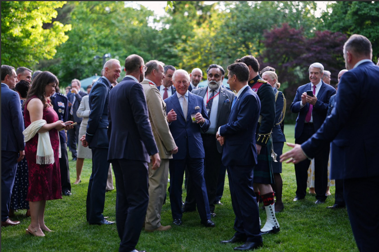 King Charles is seen smiling while standing in a garden surrounded by people wanting to speak to him 
