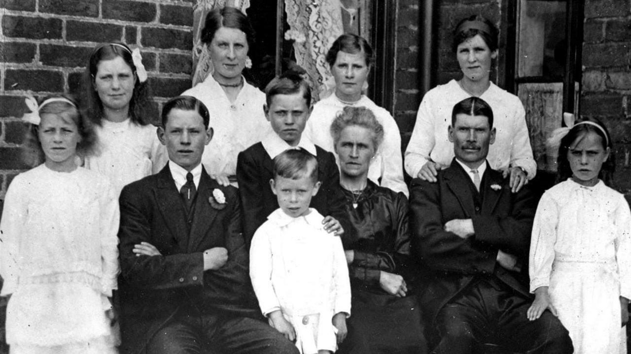 A family group picture of 11, taken outside a brick house with a window with lace curtains in the background. The picture was taken around 1918, so all the people are wearing Edwardian style formal clothing, suits for the men and lace dresses for the girls. There is also a woman wearing a dark silk dress. Three of the family, a woman and man who appear to be middle aged, and a younger man are seated, and the rest of the family, all children and young people are standing to their side or behind them. One young woman has her hands resting on the older man's shoulders. The youngest child, a boy of 6, is standing next to the older woman wearing a white sailor suit style outfit. The boy behind him has his hand on his shoulder.