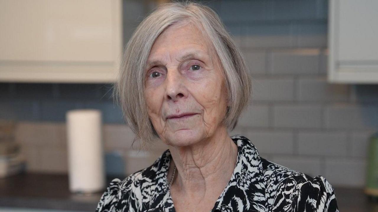 Dorothy looking straight at the camera. She has a bob hairsytyle with thin grey hair. She is in a kitchen and is wearing a black and white blouse with a flower pattern.