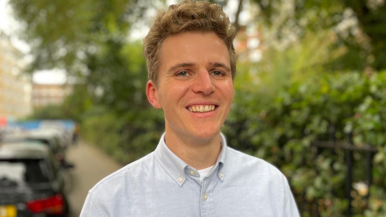 Jack May-Davis is smiling and is wearing a pale blue shirt. He is standing on a street with green hedges and a row of cars blurred in the background
