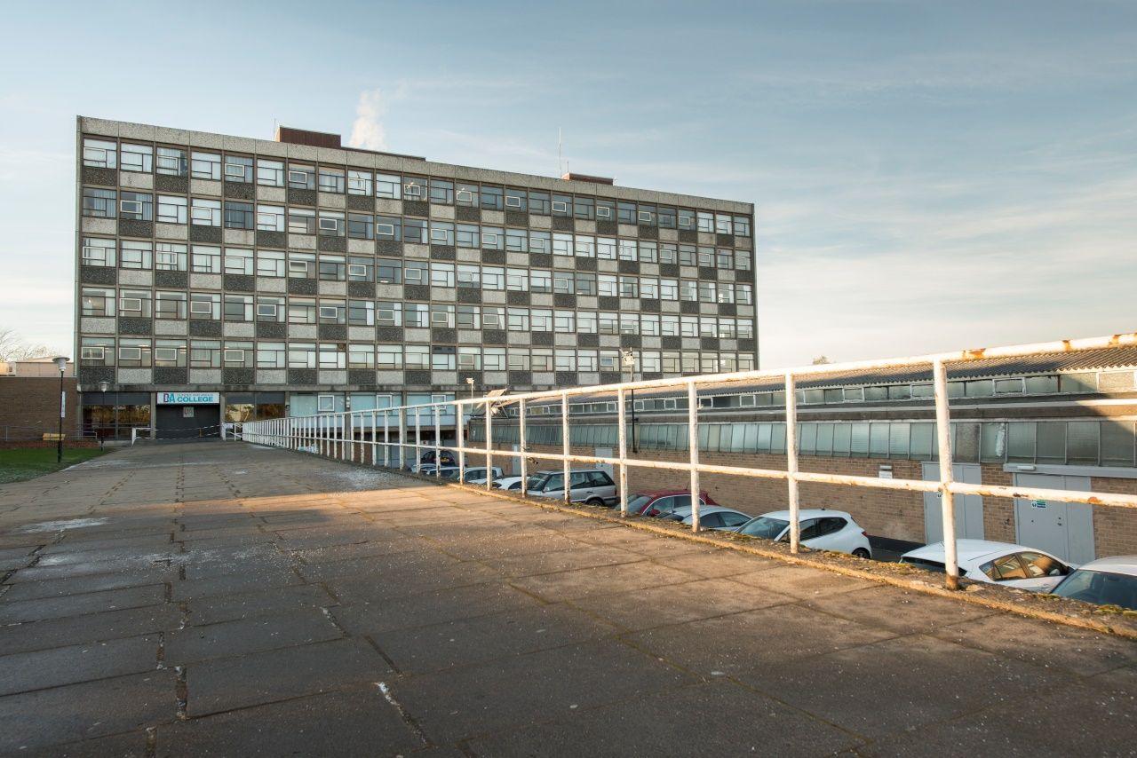 A large six-storey building set behind a paved walkway and a car park