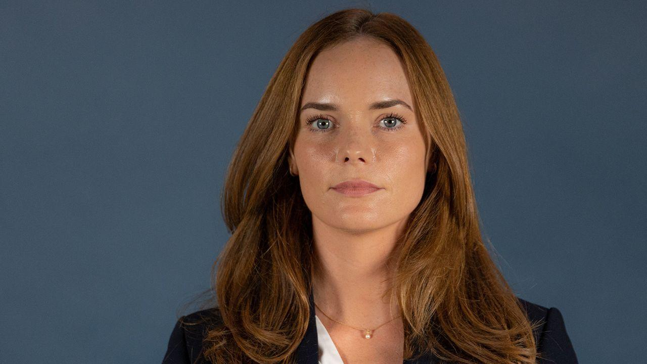 Eleanor Lyons head and shoulder portrait as she looks into the camera while wearing a suit with a necklace round her neck and straight fair hair, against a plain blue background