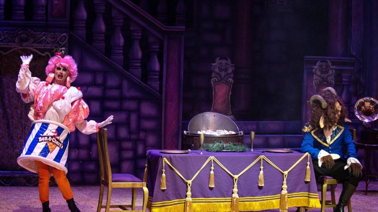 A theatre stage with two performers. On the left side, one is dressed in a flamboyant costume with bright pink sleeves, orange trousers and a skirt strapped around the waist with the text “Ben & Cherry's” on it. The performer is wearing a pink wig with dangly, ice cream-themed earrings and is standing with one arm raised and the other bent outward.
On the right side, another performer is seated at the table wearing a blue coat with gold trim and a large headpiece resembling a beast’s head with curled horns and fur.
