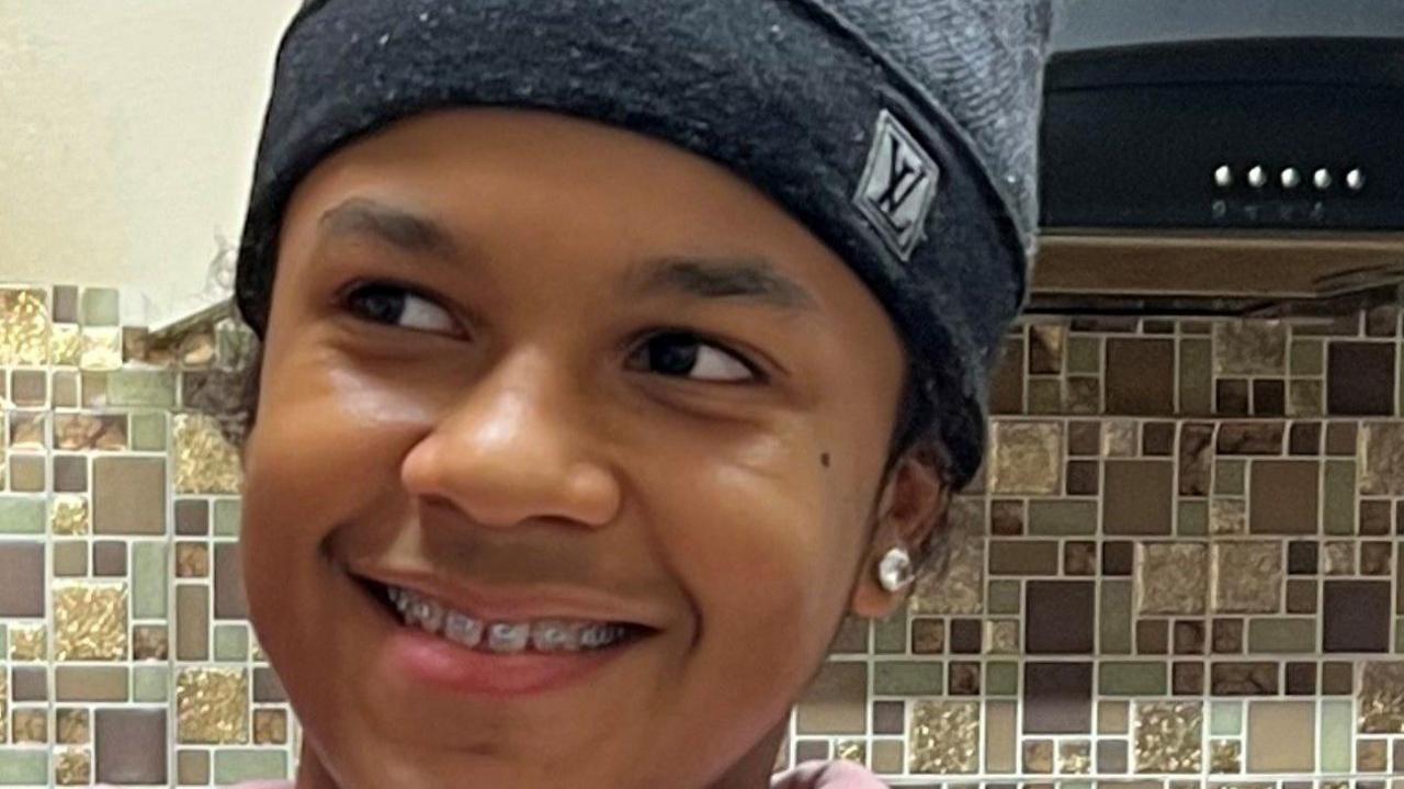 A close up picture of Leonardo who has braces and is smiling, looking away from the camera. He is wearing a Louis Vuitton grey beanie hat and has a large stud in his right ear. 