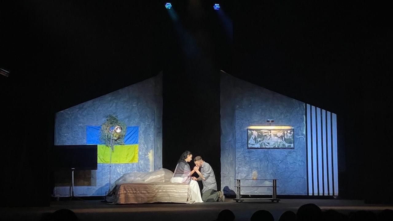 A stage set showing the cross-section of a bedroom. Centre stage a woman is sitting on a bed and a man dressed in a military uniform is kneeling in front of her and kissing her hand. On the the left wall is the Ukrainian flag, on the right wall a print of Picasso's Guernica. 