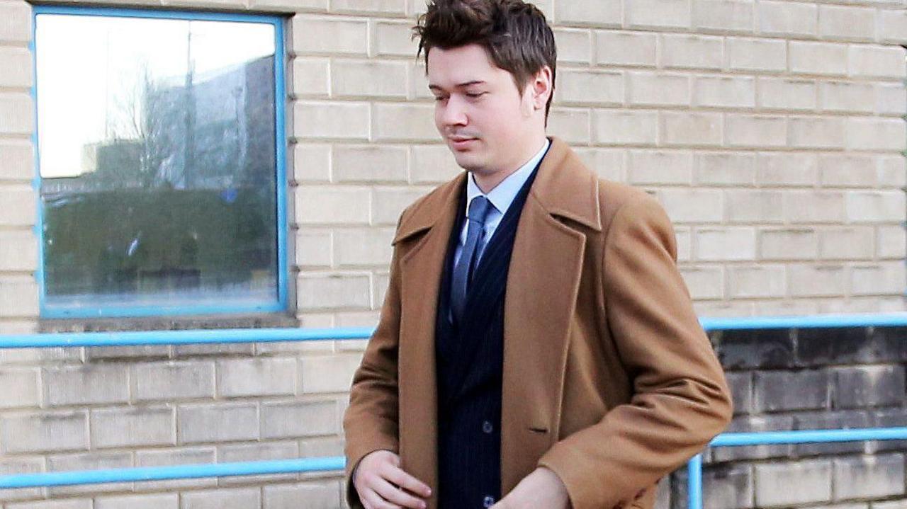 A man with short dark hair walk along a street with Wolverhampton Crown Court behind him. He is wearing a dark blue pinstripe suit and brown overcoat.