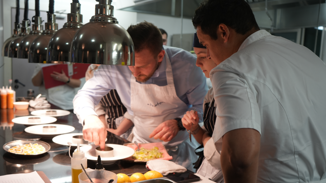 Image shows a chef teaching students how to create a dish. They are in a college restaurant. The students are crouching over a work surface as they watch an expert perfect a dish. The expert is a white man, with brown hair and is wearing a shirt and apron. He is putting his finger in a small ramekin. Six silver food heat lamps are above the work surface.