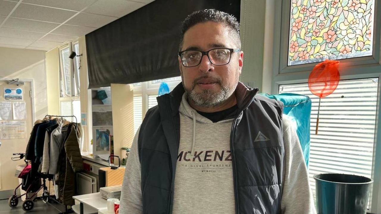 A man wearing a grey jumper and a navy gilet stood in a community centre. he wears black-rimmed glasses and has tight curled black hair. Behind him are windows with blinds on them, a coat rail and a white table.