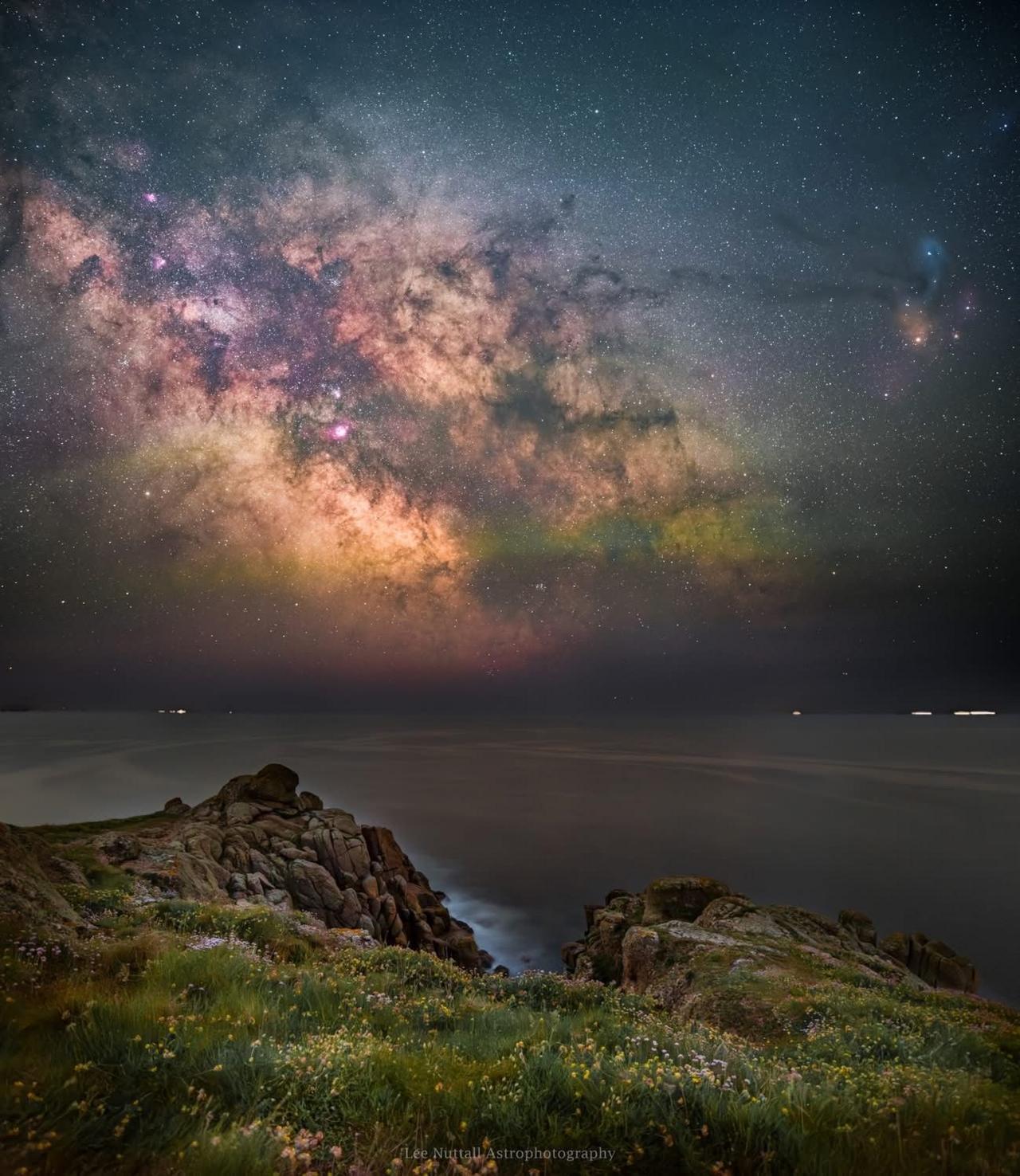 A view looking out to sea from a clifftop, with the Milky Way stretching up from the horizon.