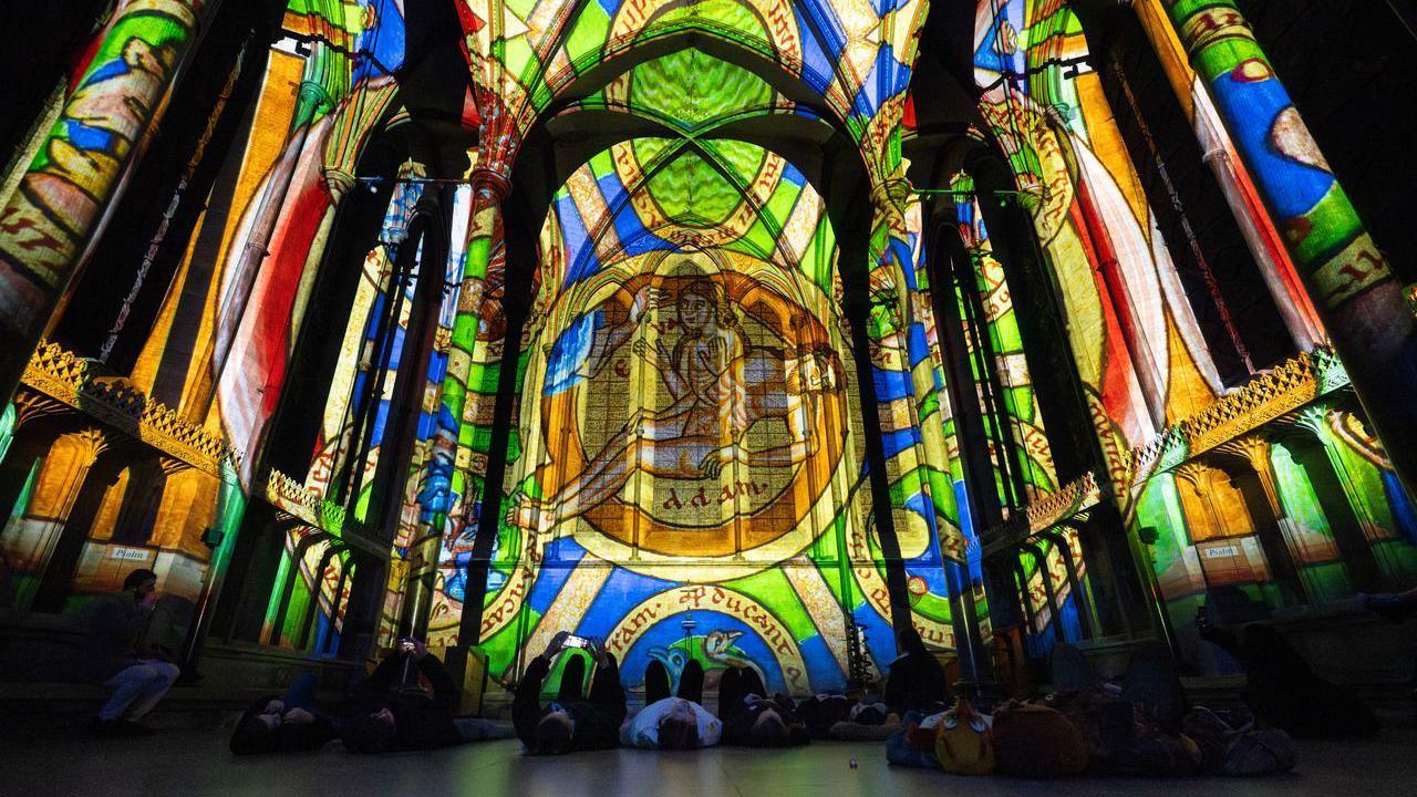 The interior of Salisbury Cathedral lit up for the Sarum Lights. A colourful medieval illustration is being projected onto a wall and the ceiling. A golden circle containing two human forms is clearly visible on the wall. Barely visible are the people lying down on the floor looking up at the patterns projected onto the wall and ceiling.