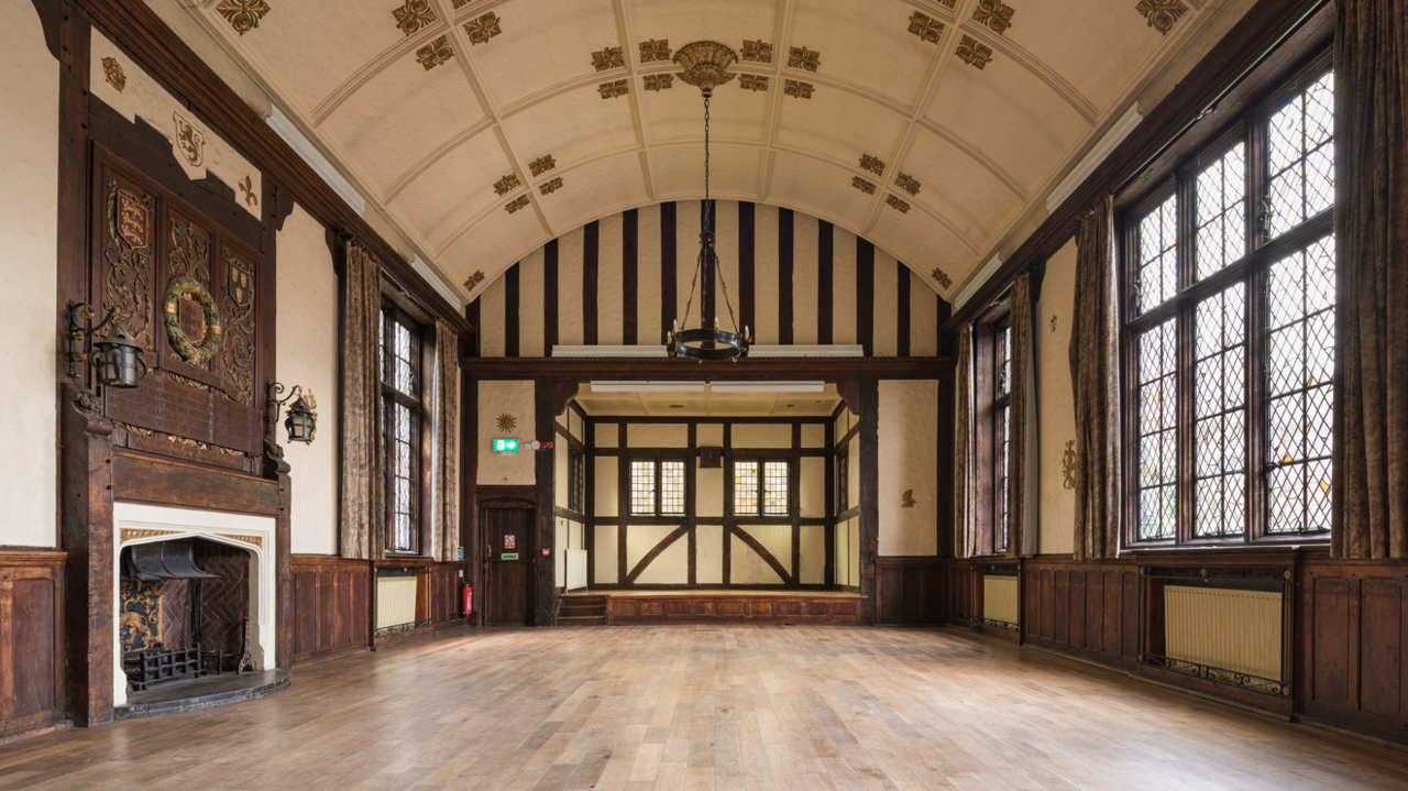 A large, empty hall. It has wooden flooring and the walls feature dark wooden panelling. To the left is a fireplace and to the right are two large windows.