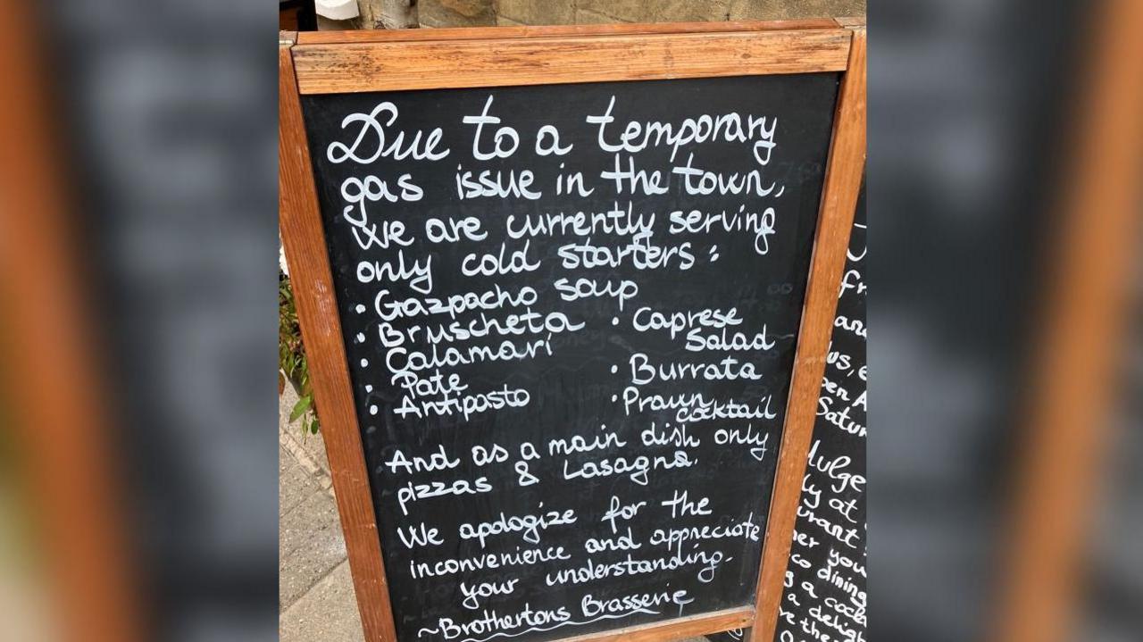 A sandwich board with chalk writing on it, which reads: "Due to a temporary gas issue in the town, we are currently serving only cold starters. Gazpacho soup, bruschetta, calamari, caprese salad, pate, antipasto, burrata, prawn cocktail. And as a main dish only pizzas and lasagne. We apologise for the inconvenience and appreciate your understanding. Brothertons Brasserie."