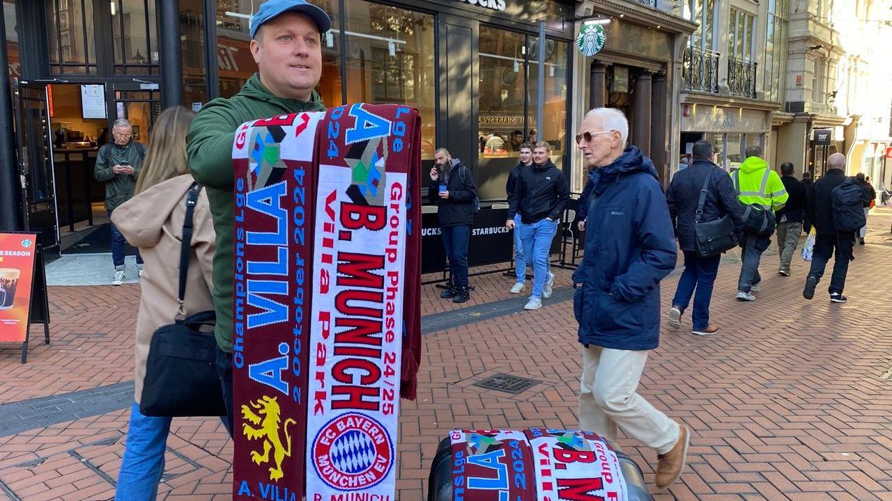 A man is wearing a blue cap holding scarfs that he is selling, that say 'A. Villa' and 'B.Munich'.