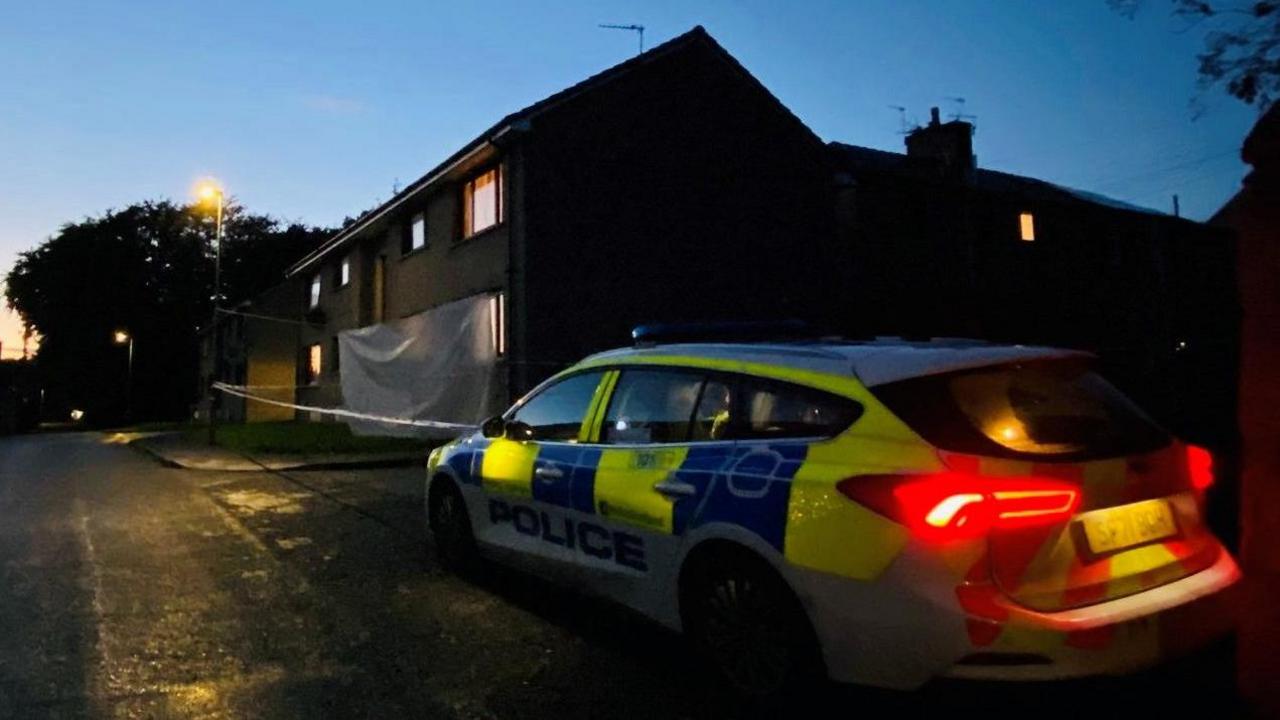 Police car with lights on outside a house, and police tape.