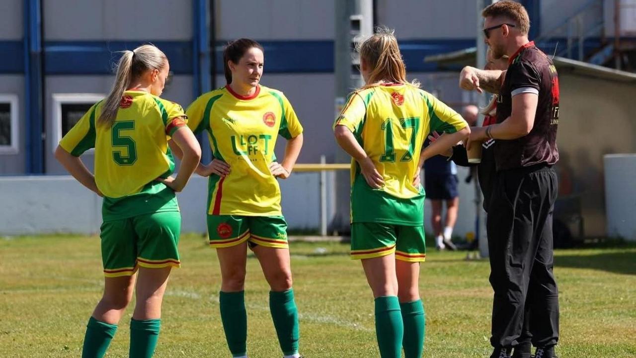 A few team members of the women's football club stood in their kit with hands on hips in talks with their manager. They have yellow shirts and green shorts, there are three players and a man with black tracksuit bottoms and a black t-shirt on.
