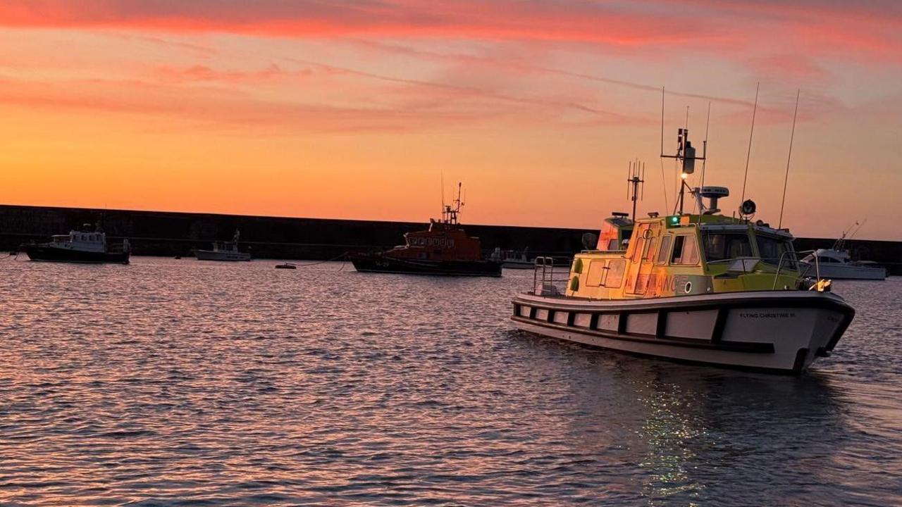 The ambulance boat on the water in Alderney at sunset, with other boats in the background, amidst pink and orange skies.