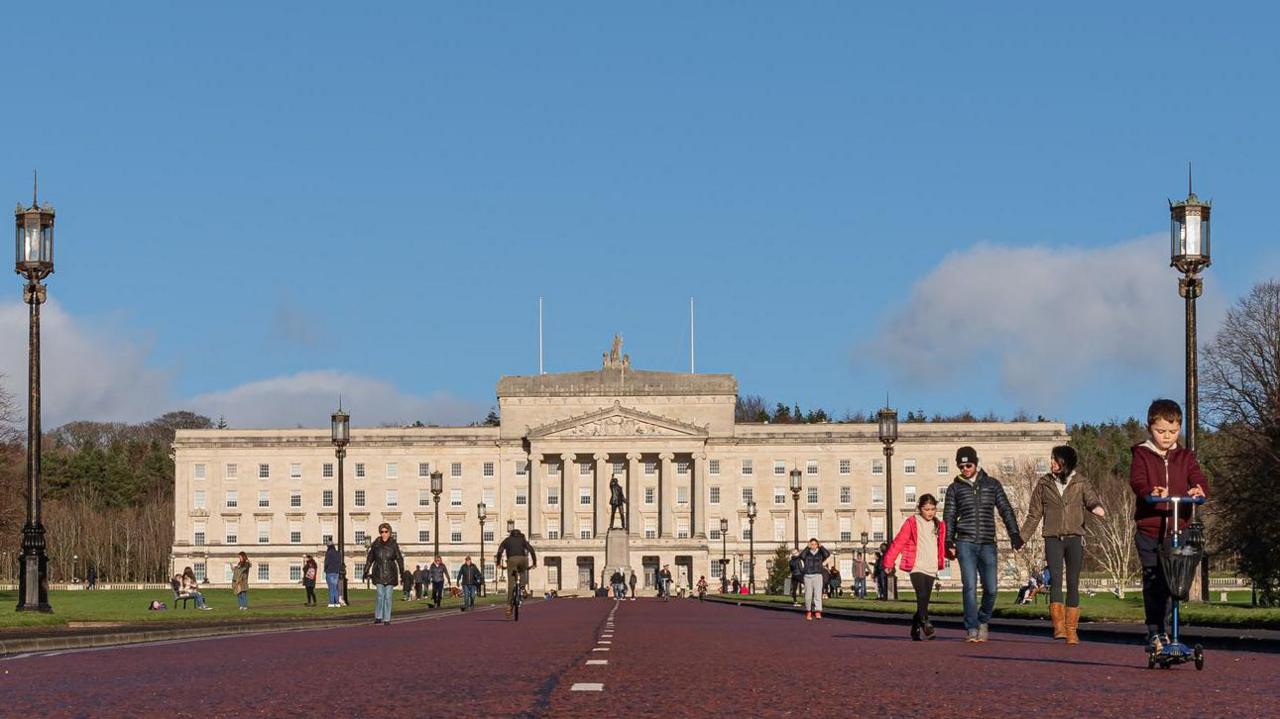 Numerous people walking, scooting, cycling and playing in front of Stormont. It is a large stone building with stairs leading up to it. It is rectangular shape and has four rows of windows along the front, and a stone turrets at the front. On top of the building is a metal statue. Dotted around the grounds are black lampposts and benches for people to sit on. There is a statue feature in front of the steps to the front door.