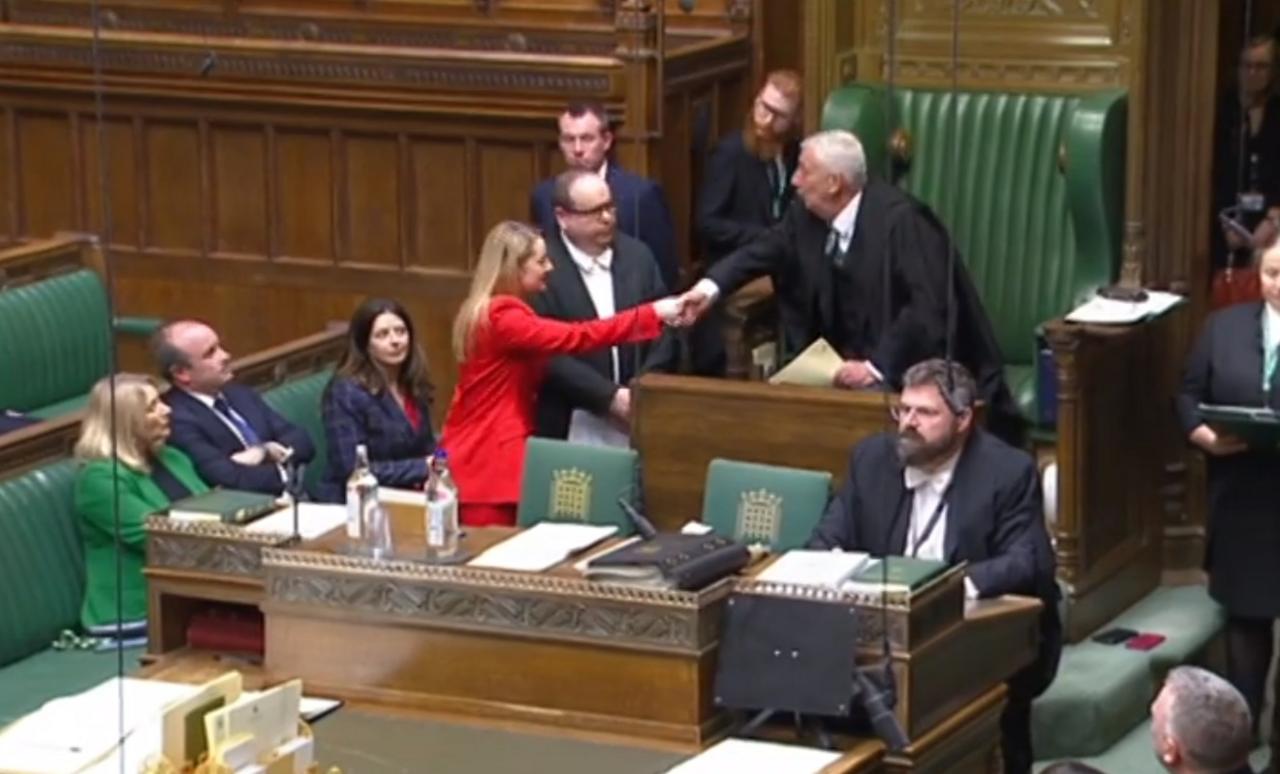 Woman in red shakes the hand of the speaker, who is standing in front of his chair in black robes