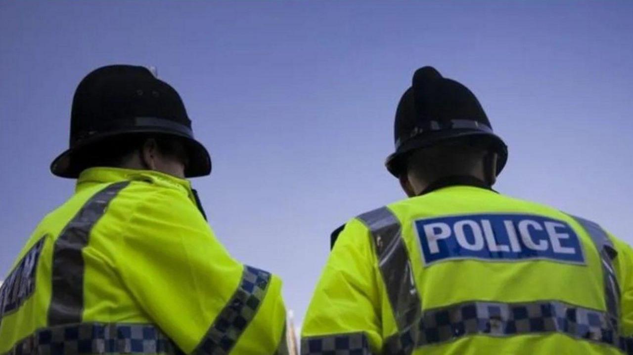 Two police officers are wearing a bright yellow jacket in this generic image. The word police is visible in full on the jacket worn by the officer on the right, in white lettering on a blue background.