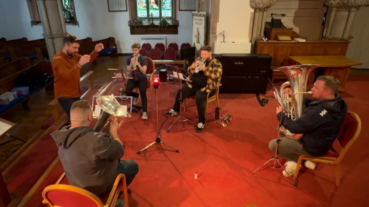 Seated musicians playing brass instruments in a church.  They are being conducted by a man standing in the centre of the group.