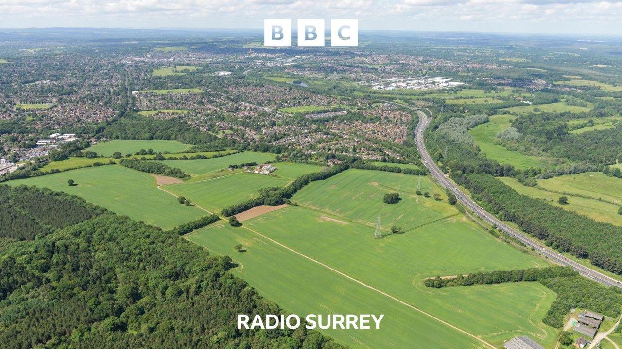 An aerial view of fields and woodland. A dual carriageway cuts across on the right and in the distance an urban area can be seen.