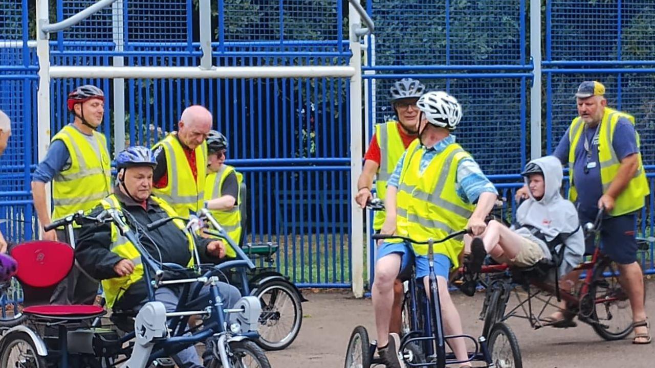 Cyclists wearing hi-vis jackets stand around inside a playground before they are about to embark on a cycle ride. The talk among each other.