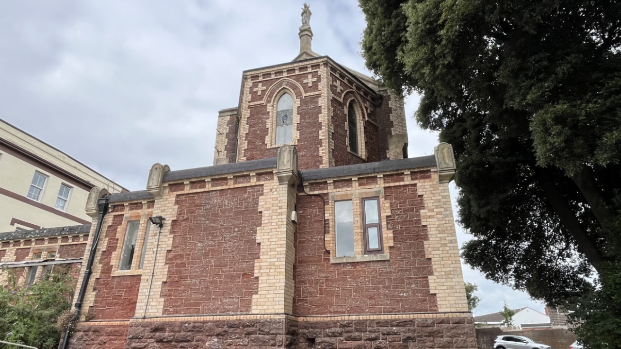 The image shows a historic red-brick and stone building with Gothic architectural features, including arched windows, ornate stone detailing, and a small tower topped by a statue.  The building is surrounded by trees and nearby structures, with a car visible in the background.