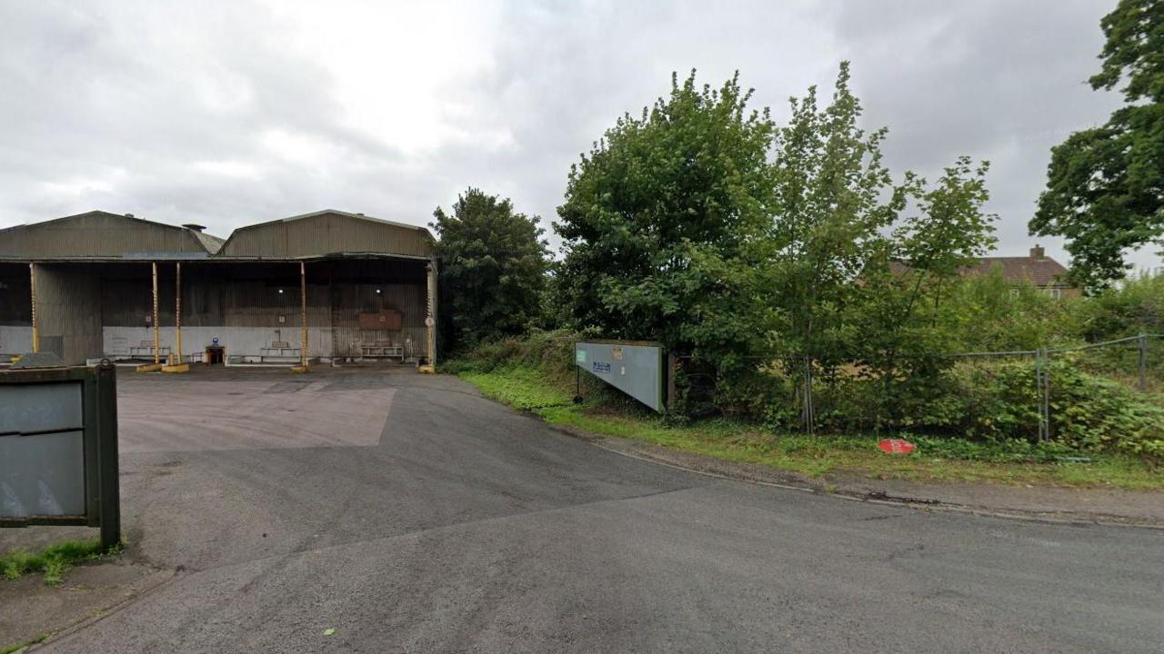 A Google maps picture of the proposed site. It shows a wide road running through an open gate leading to barn-type buildings. There are trees and shrubs at the gate.