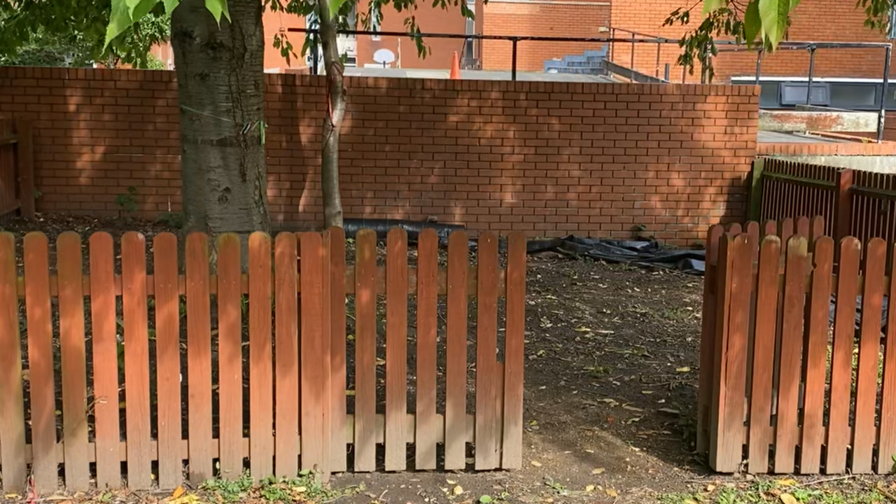 An area of soils surrounde by a fence amnd a brick wall at the back.  The area contains a single tree