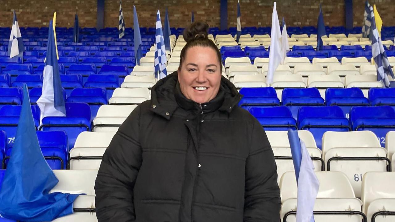 Katie Carter has dark hair tied up in a bun and is wearing a black winter coat. She is stood in front of blue and white seats which have fags placed among them.