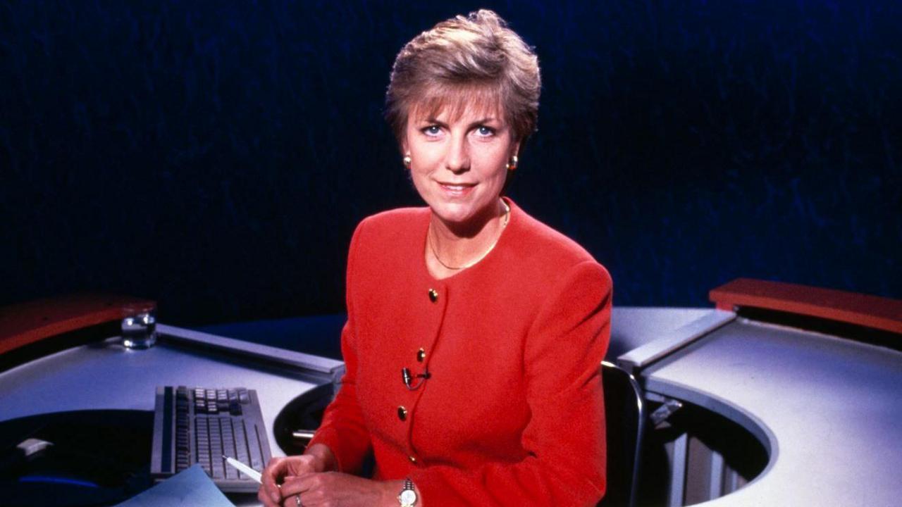 Jill Dando sitting behind a newsdesk on a television set for the 9 o'clock news in 1992. She is wearing a red suit and has short blonde hair.