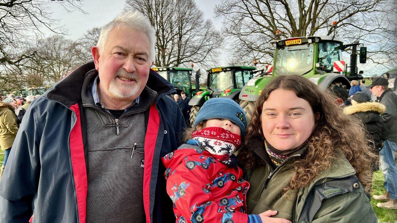 Three members of a family stand in front of a row of tractors. A man with grey hair and a goatee beard stands next to a younger woman with shoulder-length brown hair. She is holding a toddler wearing a red coat and a blue beanie hat.