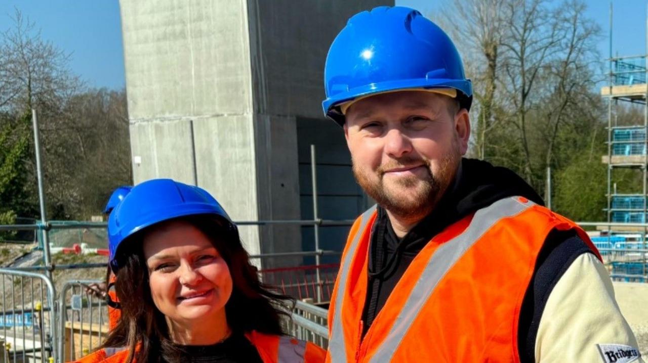Claire Rinet on the left is next to Joe Fearn and both are wearing blue hard hats and hi-vis jackets. Scaffolding is behind them in the background.