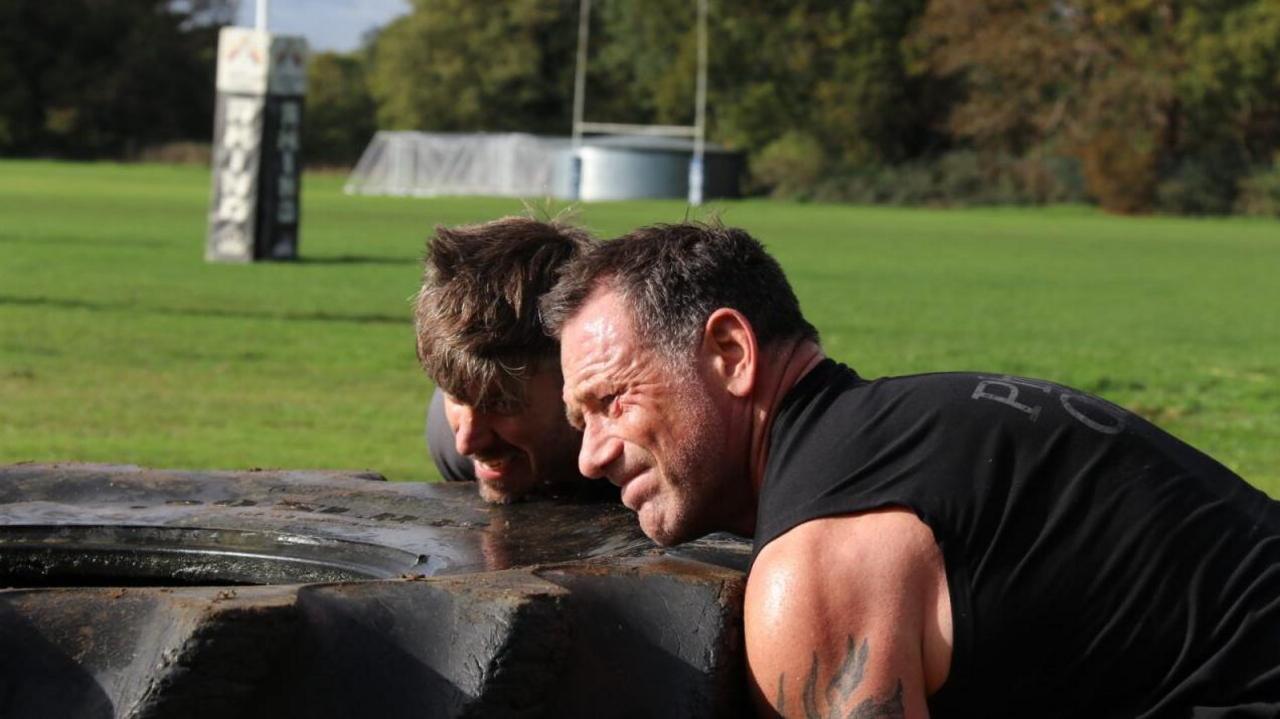 Foxy wearing a black vest and training with a friend on a green field. They are both attempting to lift a huge tyre.