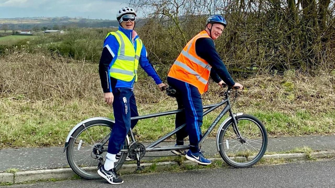 Two men sitting on a tandem bike by the side of a road. James Shone is on the back of the bike. He is wearing what looks like specialist eyewear/shades and he is smiling. The man at the front - Anthony Dutton - has his hands on the handlebars of the bike. Both men are in high vis vests and in the background hills and trees can be seen.