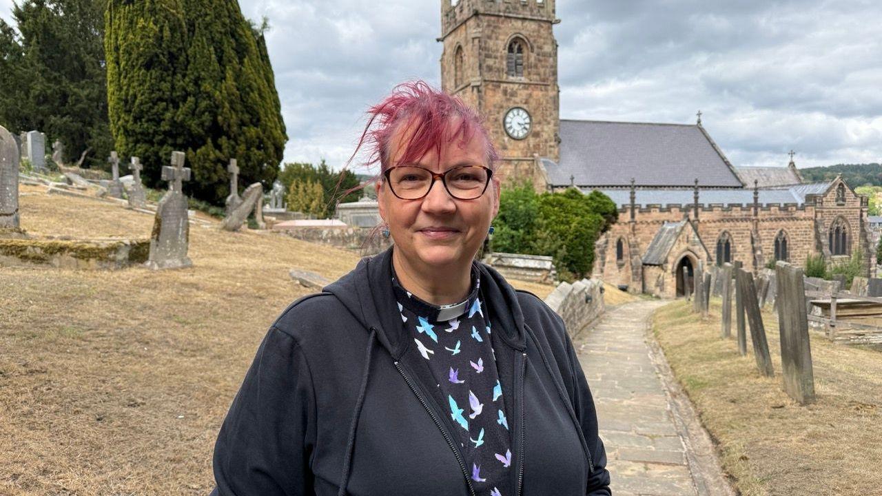 Person standing on a path in a churchyard, with gravestones all around her and a stone church in the background.
