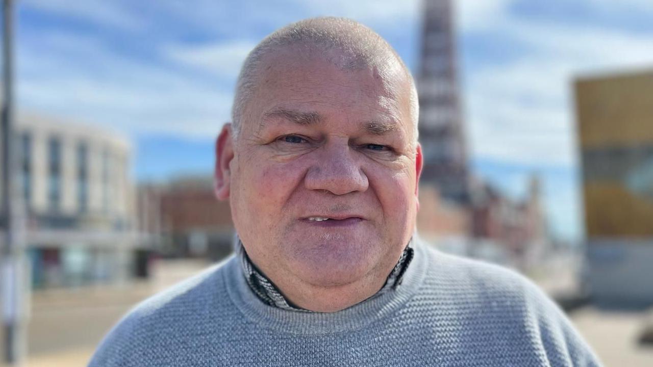 A close-up of Ian White, whose hair is shaved short. He is wearing a shirt and a blue crew-neck jumper. The blurred background features the promenade in Blackpool including the Blackpool Tower