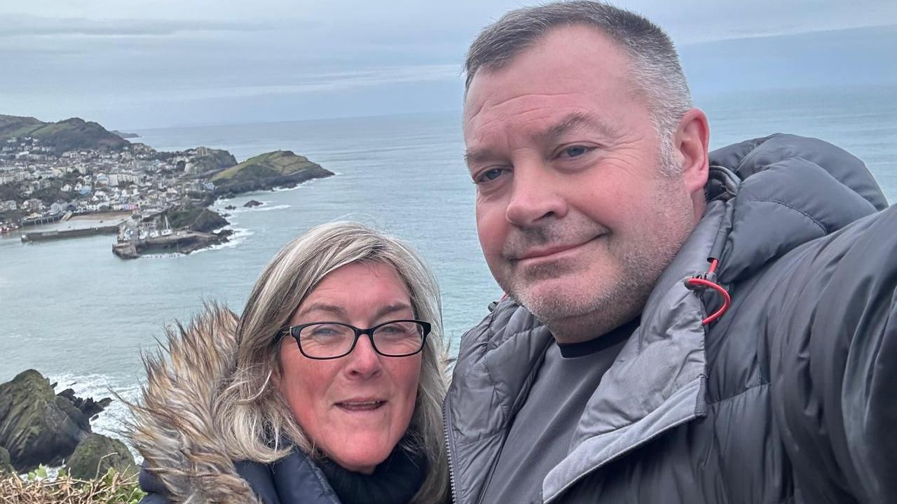 David Griffiths and Helen Moorefield dressed in outdoor clothing and standing by the sea. Both are smiling. 