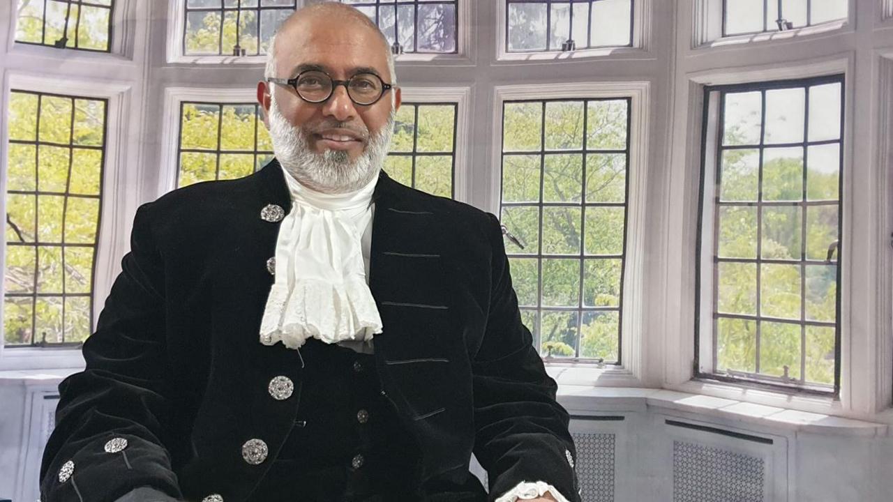 Monawar Hussain smiling for the camera. He is sitting in a room with many windows behind him. There is a desk with a few items beside him.