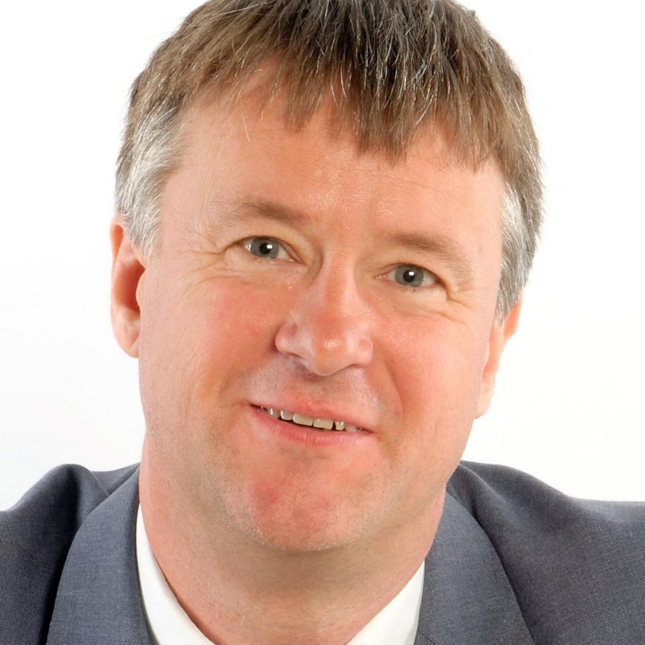 Dave has grey hair and blue eyes. He is wearing a grey blazer over a white shirt and is smiling into the camera