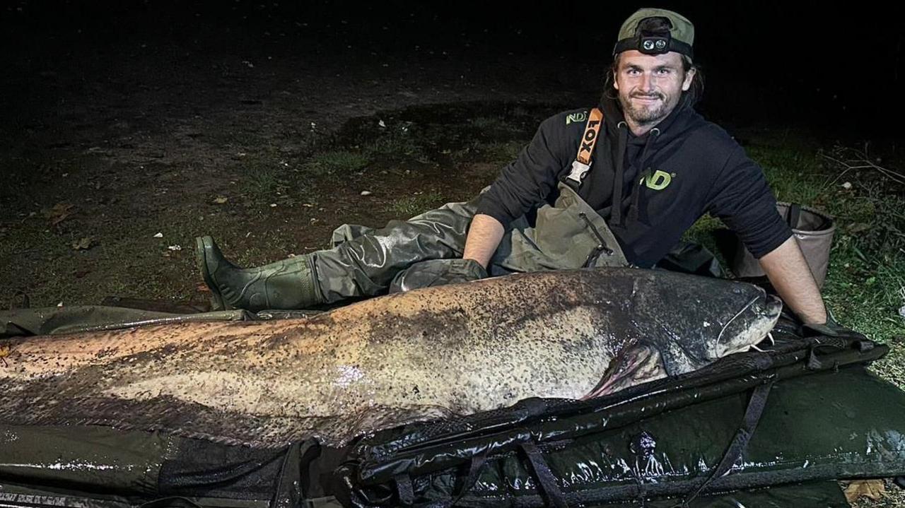 Shaun Ing, lying on the ground next to a large catfish, in a night-time image taken using a flash.