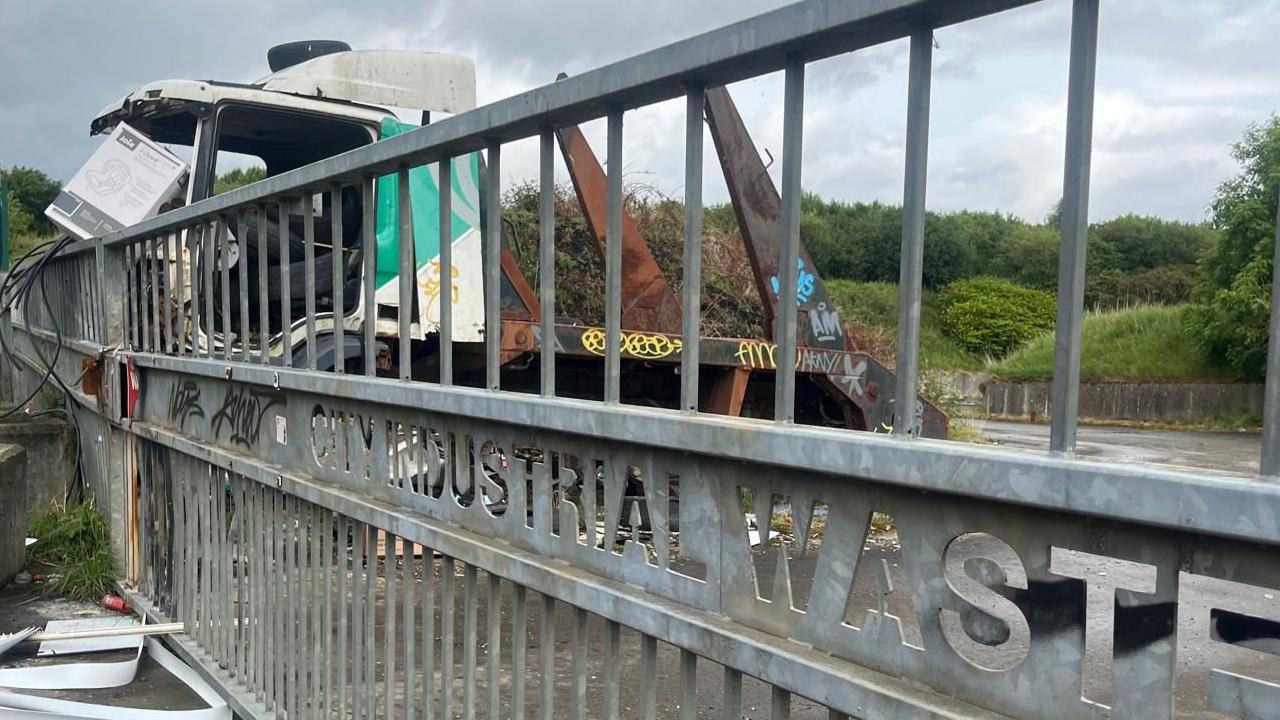 A lorry behind a fence. There is graffiti on the fence and the lorry.