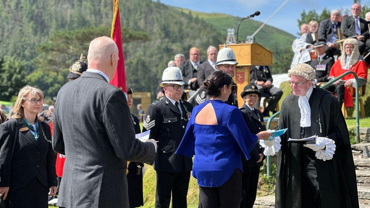 A woman in a blue top with flowing sleeves hands a petition in a blue plastic pocket to the Clerk of Tynwald, who is wearing a black robe and white wig. A man in a grey suit waits behind with his document. People in formal wear, including police in formal dress, line the area at the foot of Tynwald Hill.
