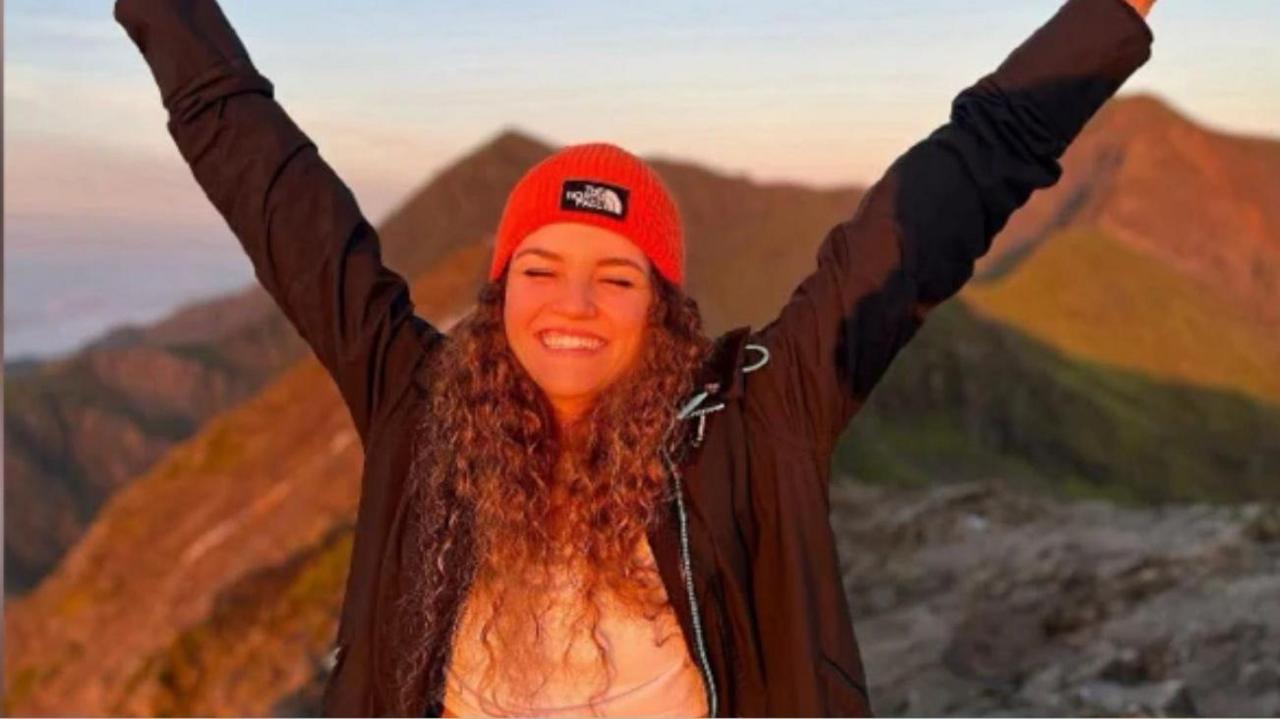 Maria Eftimova standing in the mountains, wearing a red hat and fleece, smiling at the camera and raising her hands in the air above her head.