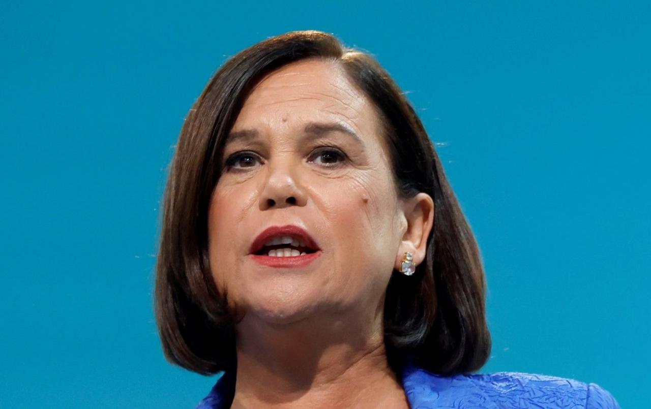 Mary Lou McDonald pictured making a speech. She is wearing a blue top and against a light blue backdrop. She has a diamond earring in her left ear