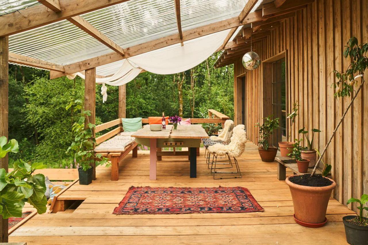 An exterior shot of a sitting area just on the porch of the home. There is a wooden bench built into the structure of the home with a table and fluffy chairs in front of it. There are plant pots surrounding the area and little discoball hangs above it. A roof made of wooden beams and plastic covering shelters the sitting area.